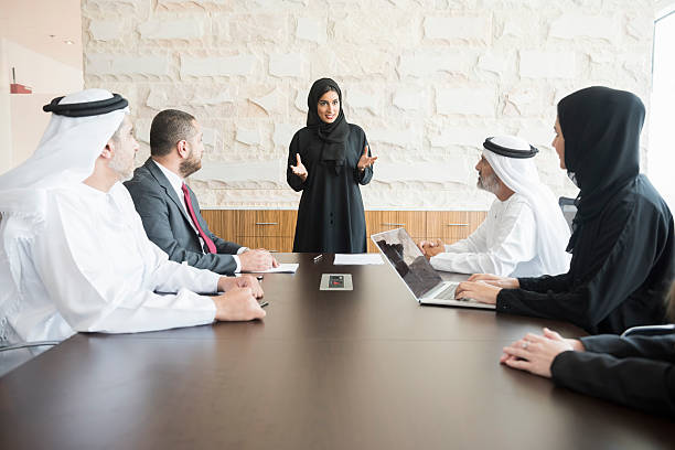 A photo of young and smiling Arab businesswoman gesturing while giving presentation. Emirati business people are wearing traditional clothes of those and abaya. Multi-ethnic colleagues are listening to her very carefully. All are in brightly lit office.